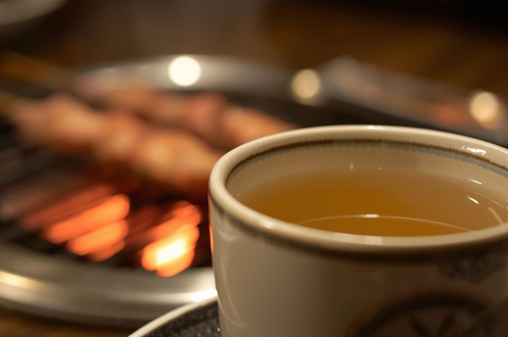 A close-up, soft-focus shot of a ceramic Japanese teacup filled with warm tea, with the blurred background showing skewers grilling over hot coals.