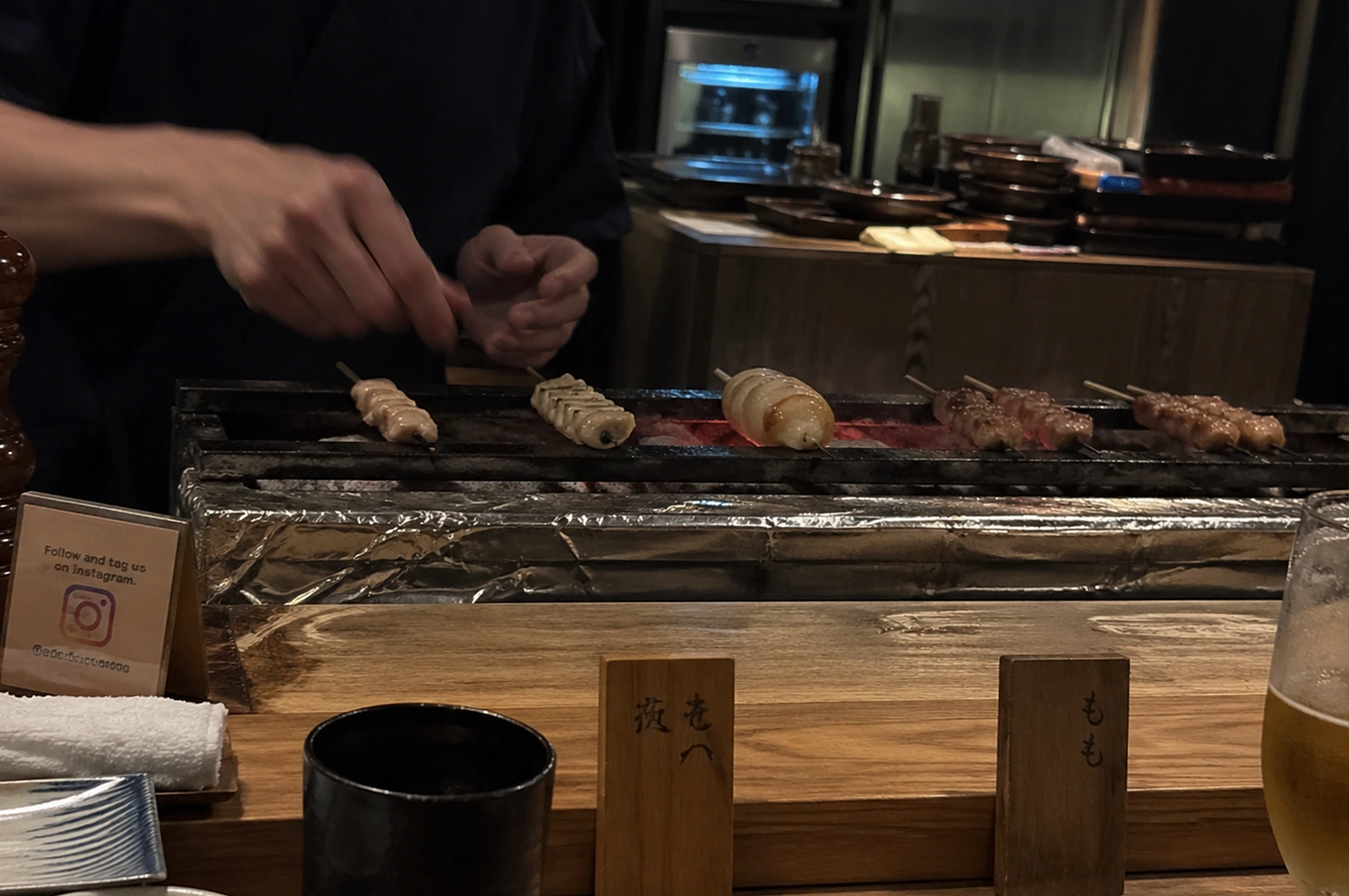 This intimate close-up shot captures the meticulous preparation process of yakitori skewers over a charcoal grill within a traditional Japanese-style izakaya. Behind the grates where five sticks of various meats—including what looks like pork belly, chicken breast, and meatballs—sizzle with rising wisps of smoke, a chef’s hands are carefully adjusting each skewer to ensure even cooking. The grill itself is lined with foil for neatness, and sits below a wooden counter where the customer's perspective begins. In the foreground, there are ceramic dishes, wooden blocks with hand-painted Japanese kanji, and an half-empty glass of amber beer, all suggesting the atmosphere is casual yet focused on high-quality grilled delicacies. An small sign requests followers on social media, integrating contemporary hospitality into the timeless culinary practice.