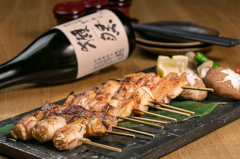 Close-up of grilled yakitori chicken skewers on a black slate board with sake bottle and mushrooms.