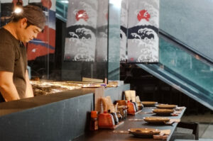 Japanese yakitori chef grilling skewers behind a counter with traditional wave and sun banners.