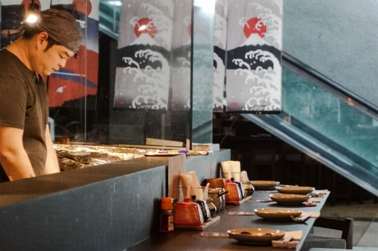Japanese yakitori chef grilling skewers behind a counter with traditional wave and sun banners.