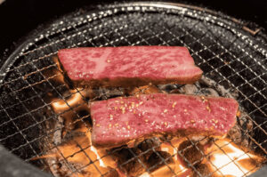 Close-up of two thick slices of premium marbled beef sizzling on a wire mesh grill over hot charcoal flames.