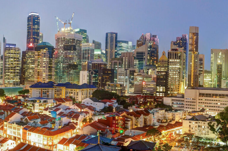 A dusk view of the Singapore skyline featuring traditional red-roofed shophouses in the foreground contrasting with towering modern CBD skyscrapers in the background.