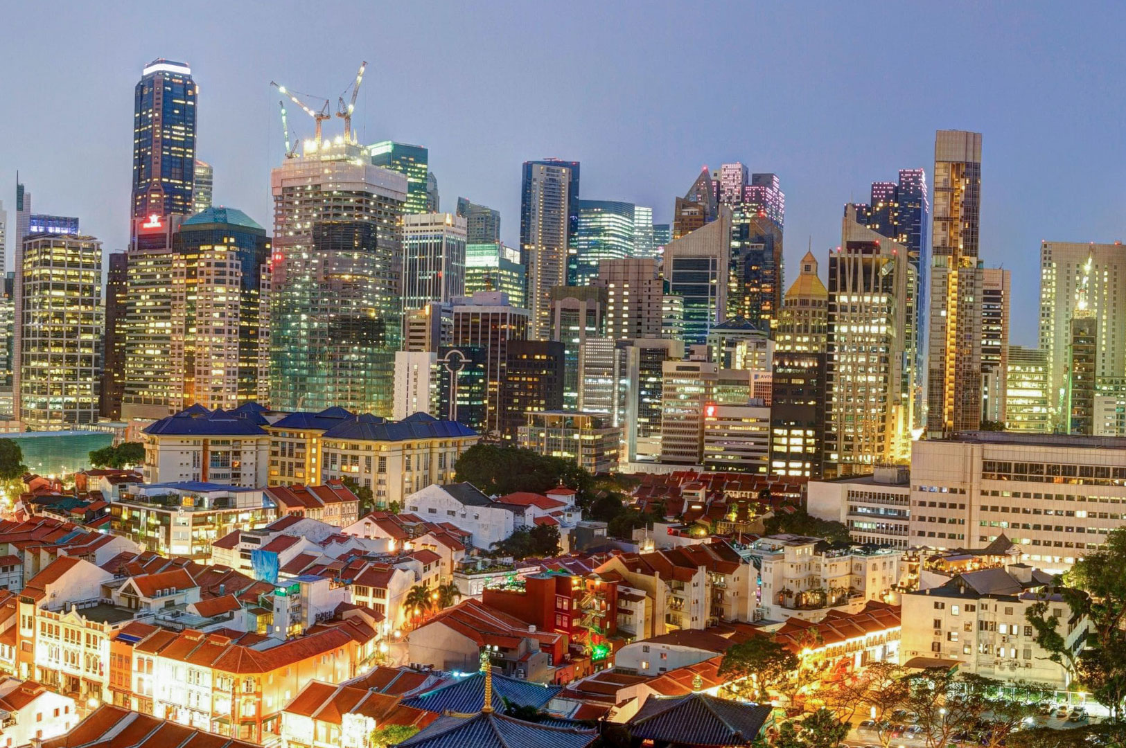 A dusk view of the Singapore skyline featuring traditional red-roofed shophouses in the foreground contrasting with towering modern CBD skyscrapers in the background.