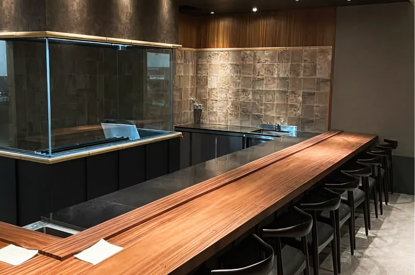 A sleek, modern Japanese-style counter with a long wooden tabletop, black minimalist chairs, and a glass-enclosed kitchen prep area.