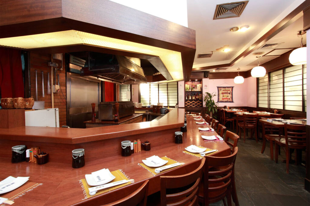 Interior view of a traditional Japanese restaurant featuring a large wooden u-shaped counter and a central robata charcoal grill station.
