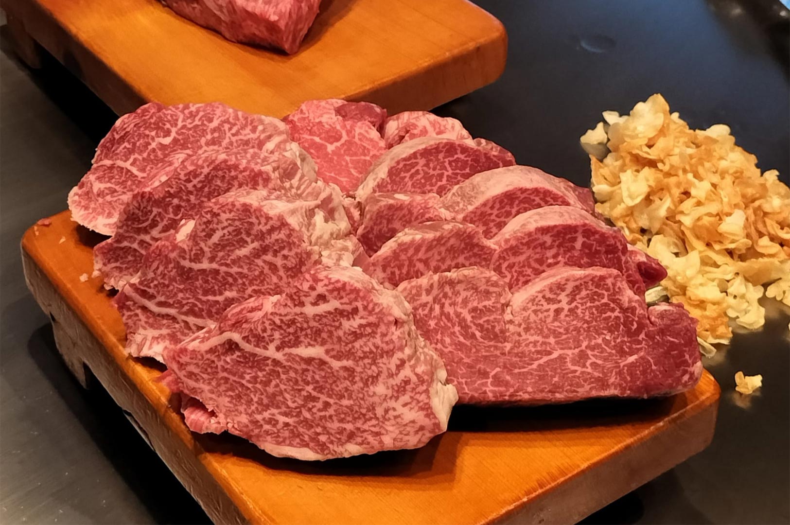 Beautifully marbled slices of raw Wagyu or high-quality beef arranged on a wooden cutting board next to a pile of crispy fried garlic chips.