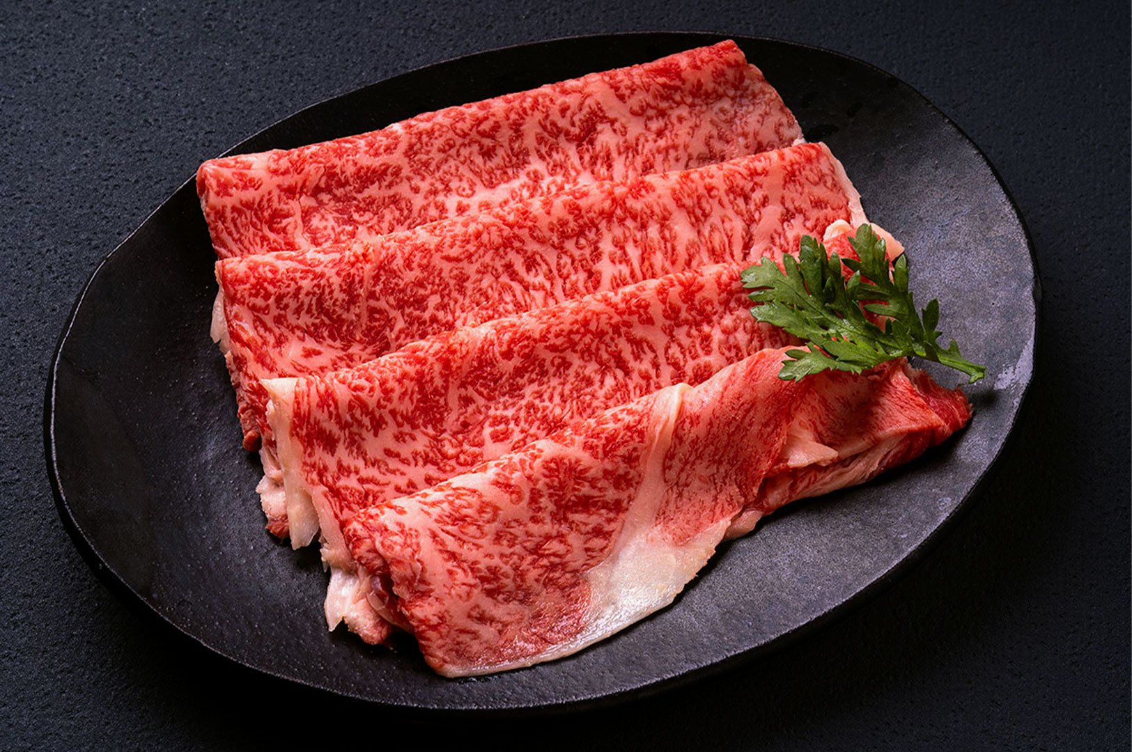 Thinly sliced raw beef with high-quality marbling folded neatly on a textured black ceramic serving plate.