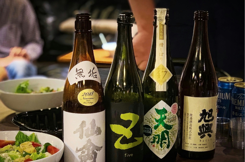 A close-up of four large bottles of Japanese sake with various traditional labels, standing on a table next to a fresh salad.