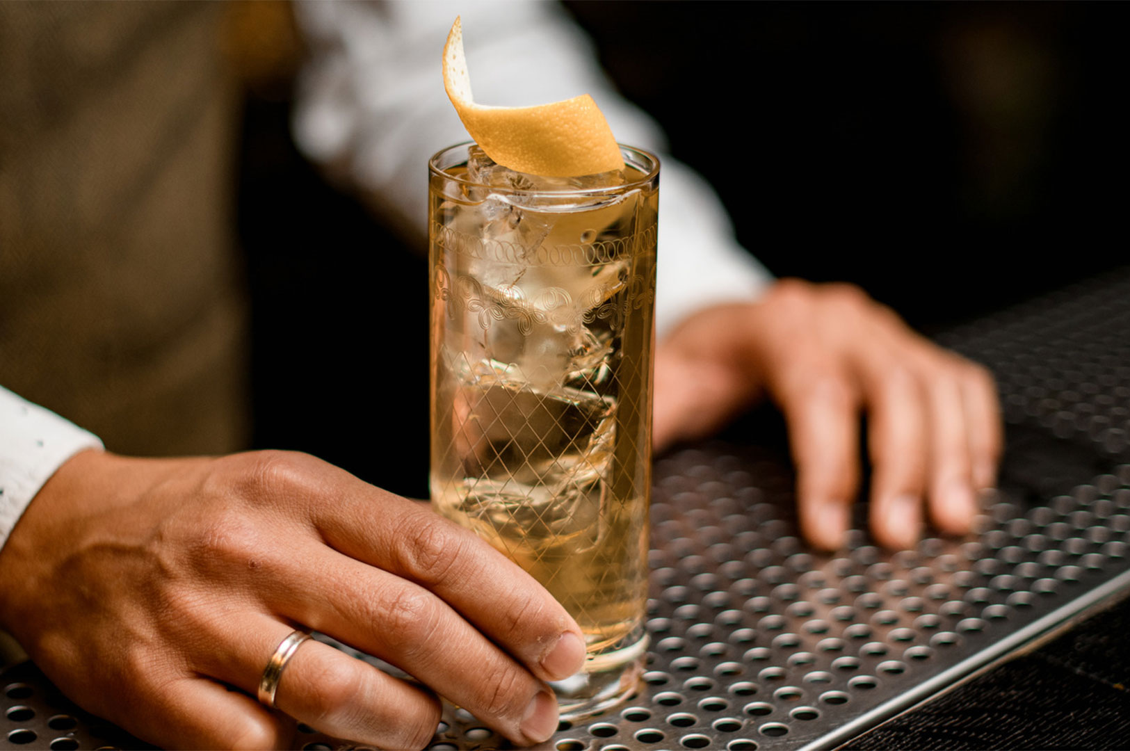 Bartender holding a tall glass highball cocktail garnished with a lemon twist on a bar counter.