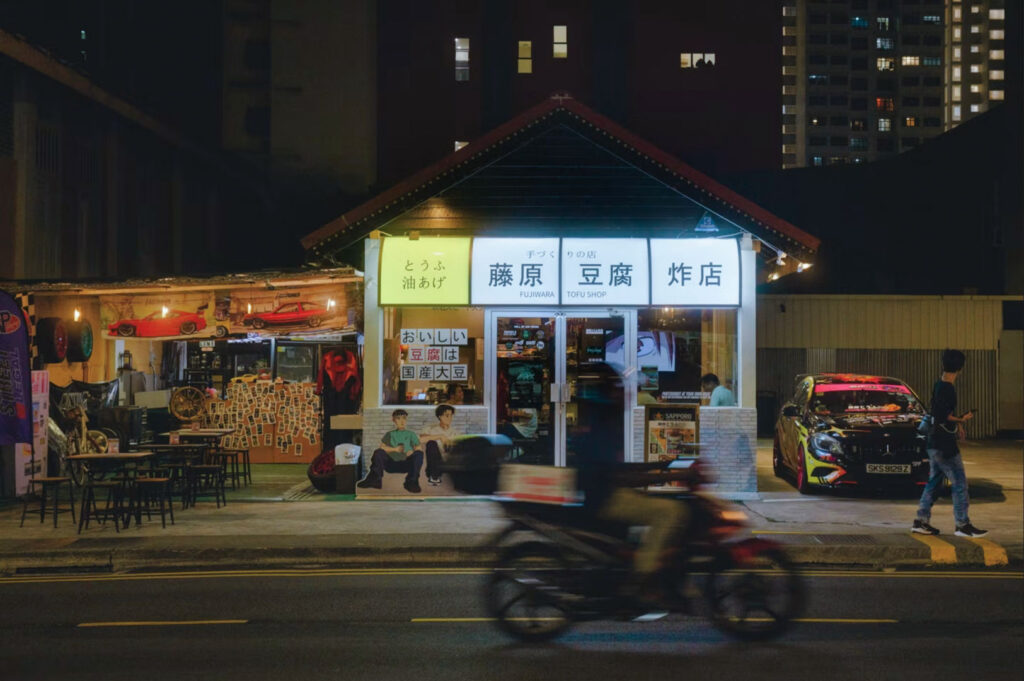 Nighttime street view of a Fujiwara Tofu Shop storefront inspired by Initial D, featuring illuminated Japanese signage, a parked car, and a motorbike passing by with motion blur.