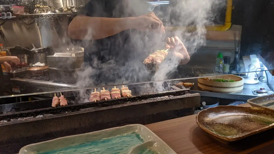 Chef grilling skewers over open flame with smoke rising in an intimate kitchen setting. Warm, rustic atmosphere with dishes ready to be served.