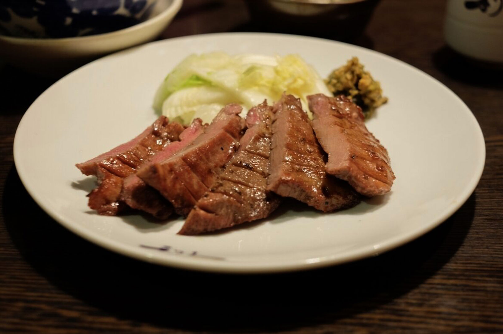Slices of grilled beef tongue (Gyutan) served on a white plate accompanied by fresh cabbage and savory condiments.