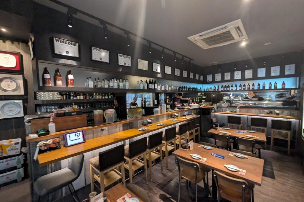 A modern izakaya interior with a long wooden bar counter, high stools, awards displayed on the wall, and rows of sake bottles.