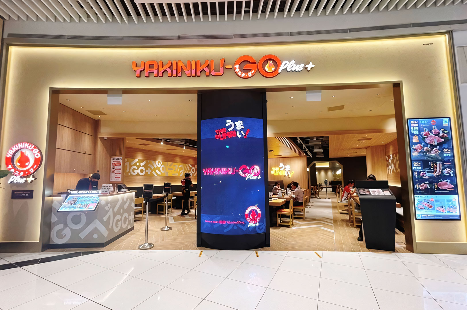 The brightly lit storefront entrance of Yakiniku-GO Plus restaurant, featuring wooden interiors and digital signage displays.