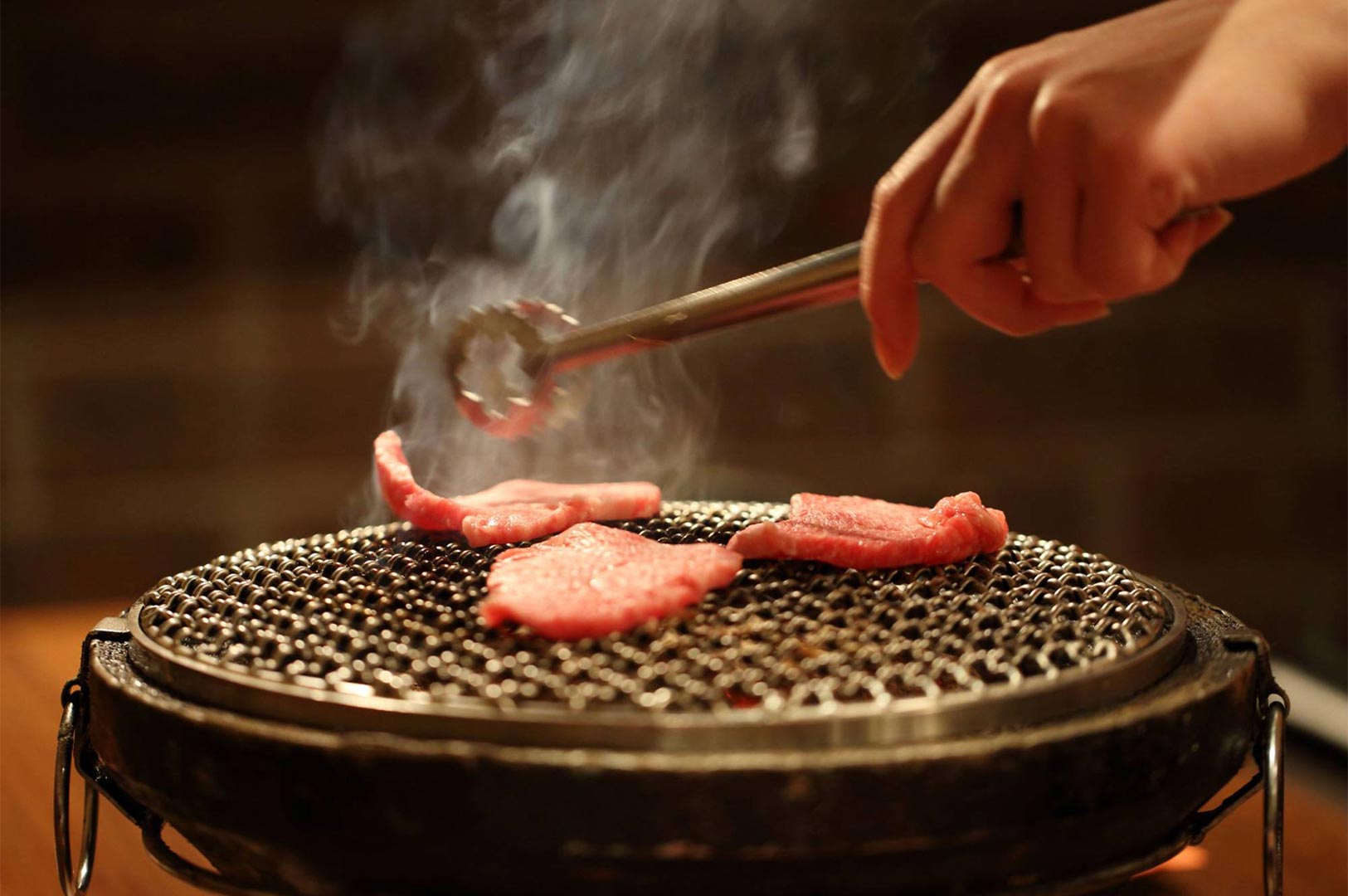 Close-up of thin slices of raw meat being grilled on a small, round wire-mesh tabletop yakiniku grill, held by metal tongs.