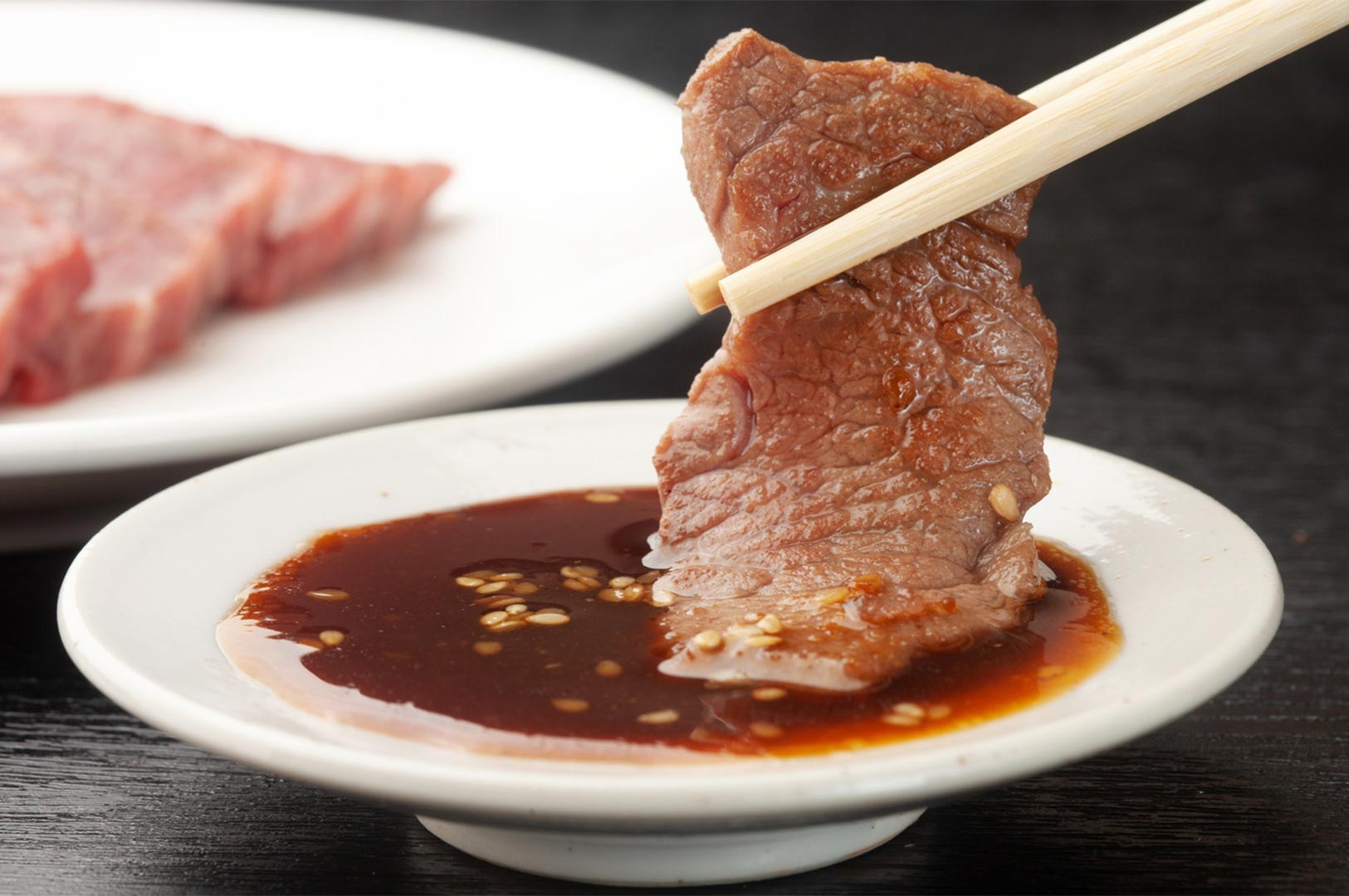 A piece of grilled, cooked meat held by wooden chopsticks being dipped into a soy-based sauce with sesame seeds in a small white dish.