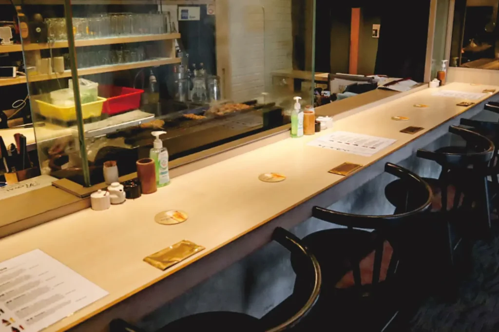 A long, empty restaurant counter with wooden chairs and a glass barrier. Condiments, menus, and sanitizer bottles are placed on the countertop, conveying a clean, welcoming atmosphere.
