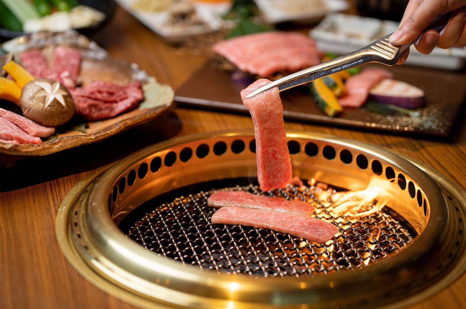 A diner using metal tongs to hold a slice of raw, marbled beef over a hot Japanese Yakiniku grill with other meats in the background.