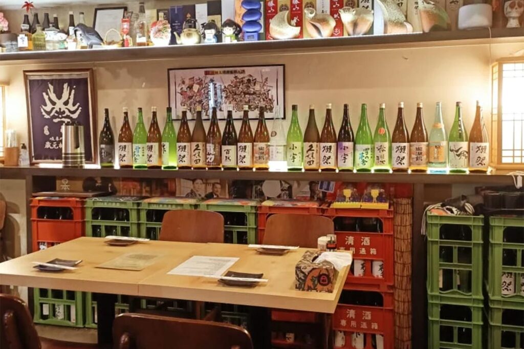 Close-up of a restaurant dining area featuring a wide variety of Japanese sake bottles displayed on a shelf above red and green crates.
