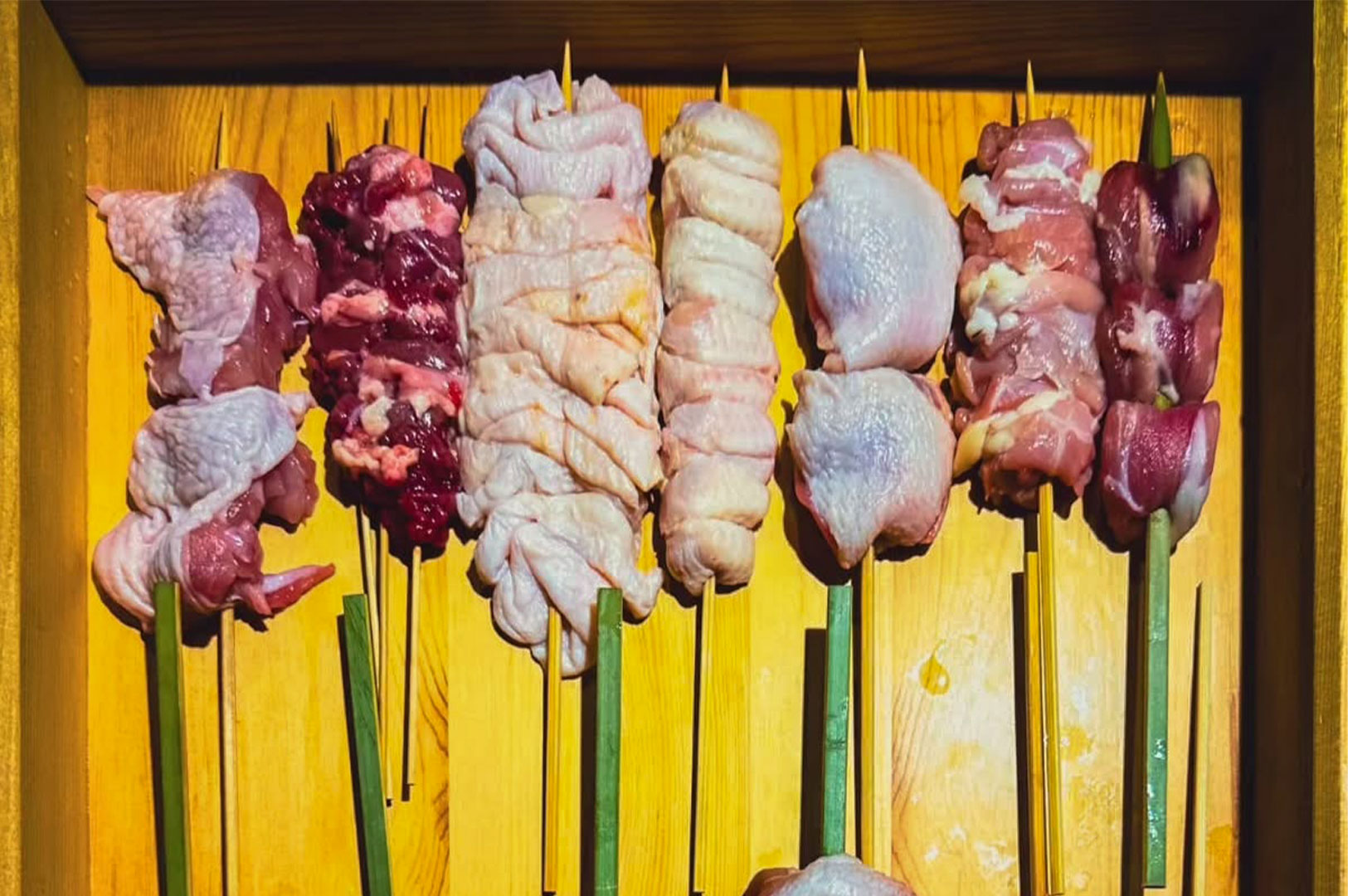 A wooden tray showcasing an assortment of raw, pre-skewered yakitori, including different cuts of chicken meat and organ meats.