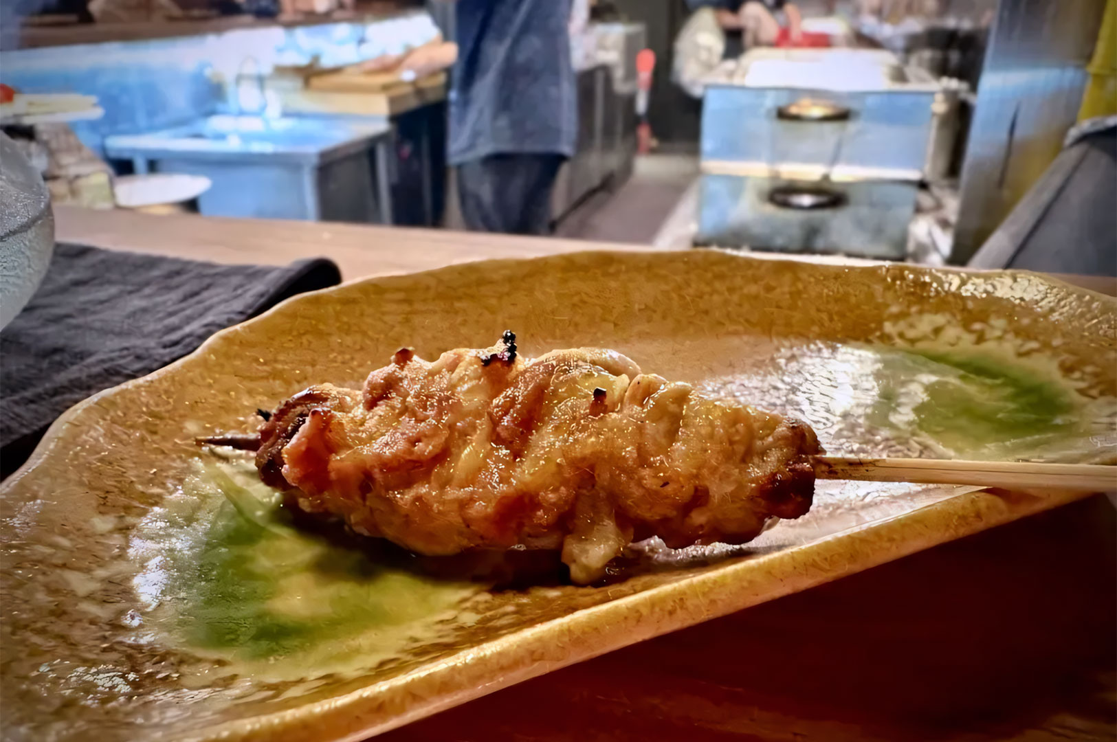 A single piece of perfectly grilled, golden-brown chicken skin or thigh yakitori skewer resting on a rustic, patterned plate.
