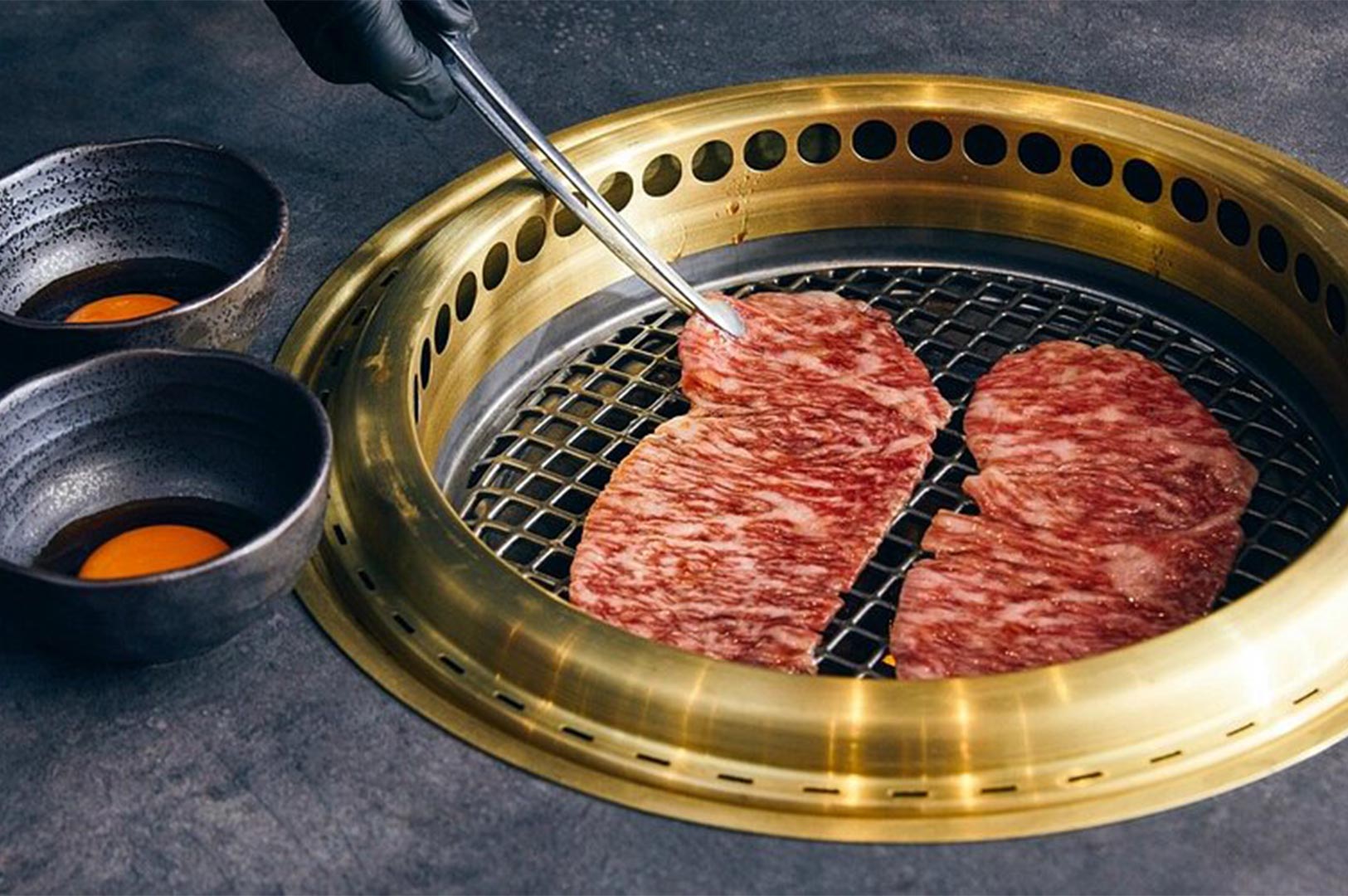 Two wide slices of highly marbled beef cooking on a wire-mesh grill set into a stylish brass or gold-toned yakiniku table grill, with dipping bowls nearby.