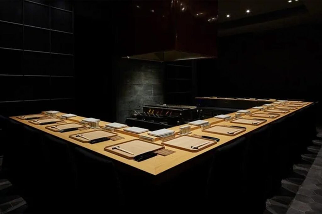 A minimalist, upscale omakase-style dining room with a light wood counter, dark walls, and dramatic spotlighting over each place setting.