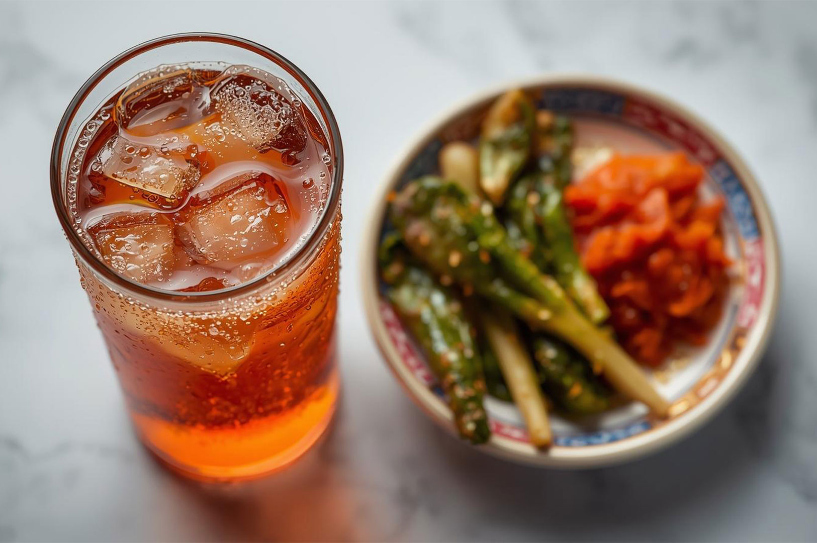A refreshing glass of iced tea served alongside a bowl of kimchi and pickled vegetable side dishes.