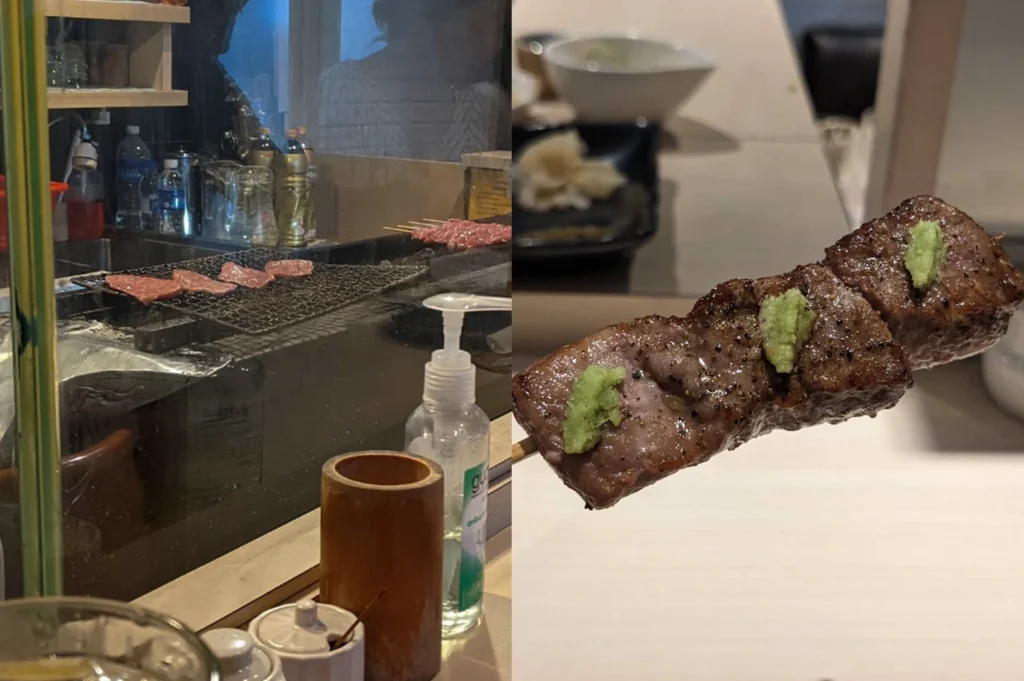 This composite image, splitting down the middle, captures a two-part culinary experience centered around yakitori. On the left, a view from inside a bustling Japanese restaurant kitchen shows four thin slices of meat grilling over hot charcoal on a wire rack, with other skewers visible further back. A clear glass partition separates the kitchen from the dining area, which features small wooden containers, condiments, and a large bottle of hand sanitizer. A blurred image of a dining patron is also visible through the window. On the right, a detailed close-up shot focuses on a single finished skewer featuring three juicy, perfectly grilled pieces of beef seasoned with black pepper and topped with generous portions of vibrant green wasabi paste. A single wooden skewer holds the meat, with the blurred background hinting at a dining table setting.