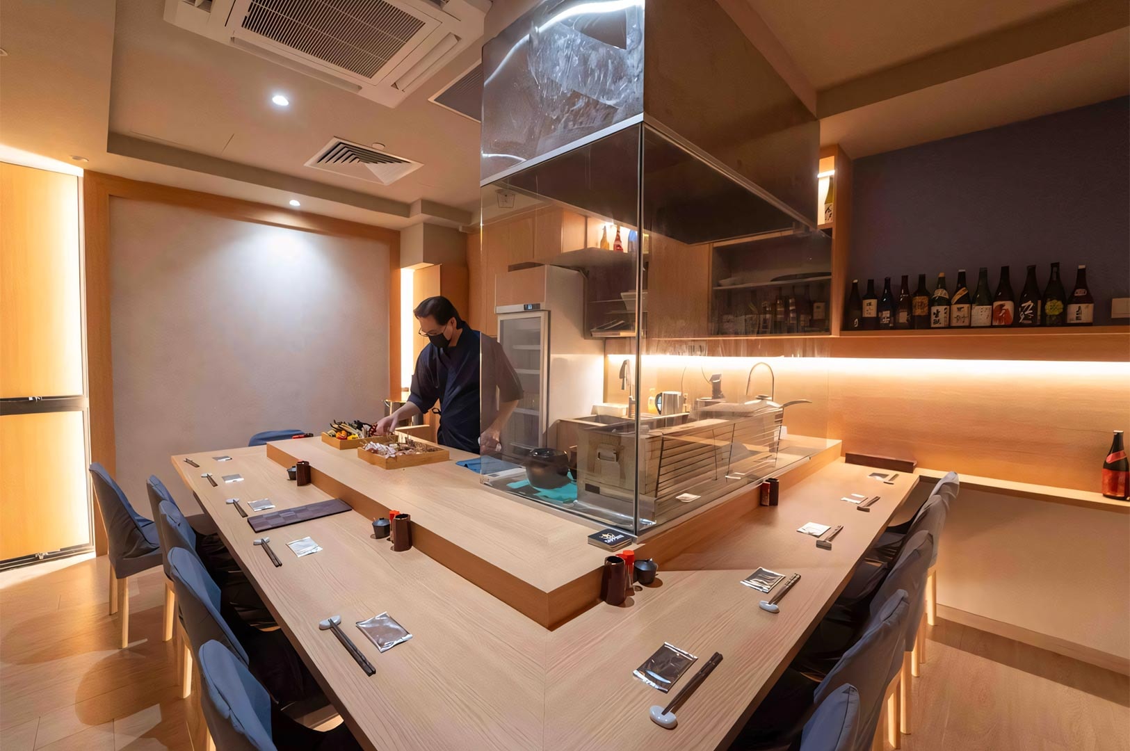 A chef preparing food inside a glass-enclosed grill station surrounded by L-shaped wooden counter seating in a modern Yakitori restaurant.