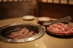 Slices of raw beef cooking on a round charcoal Yakiniku grill with a plate of fresh meat ready to be grilled in a warm restaurant setting.