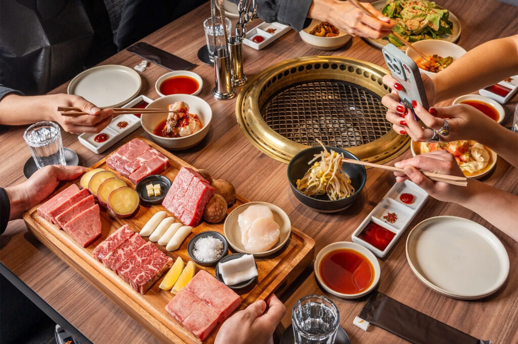 Overhead view of friends enjoying a Japanese BBQ feast, featuring a large wooden platter of premium Wagyu beef cuts, scallops, and vegetables surrounded by dipping sauces.