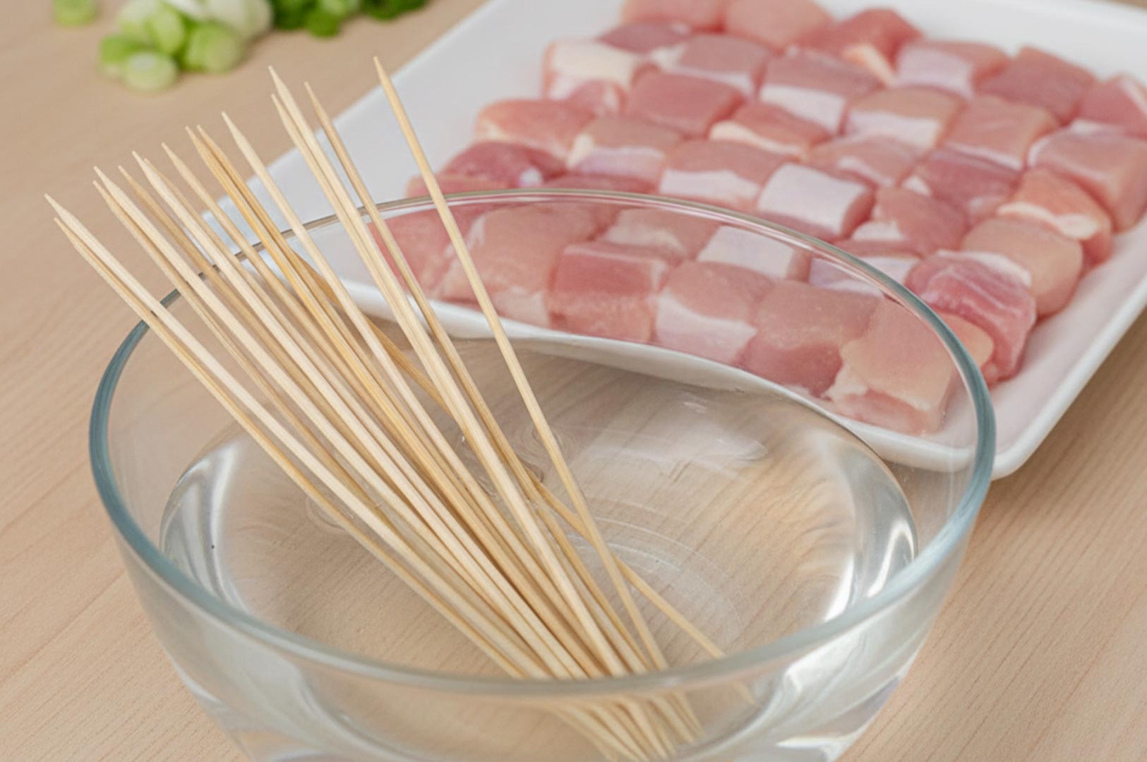 Kitchen preparation for yakitori showing bamboo skewers soaking in a bowl of water next to a plate of raw, cubed chicken thighs.