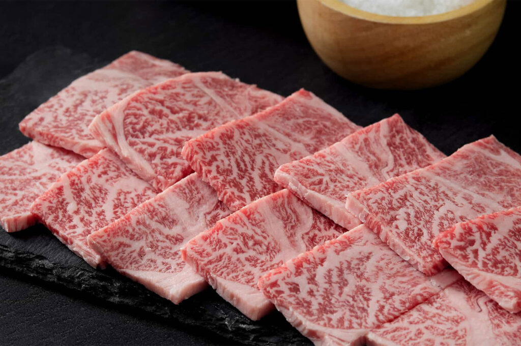 Close-up of highly marbled raw Wagyu beef slices arranged neatly on a black stone plate next to a small bowl of sea salt.