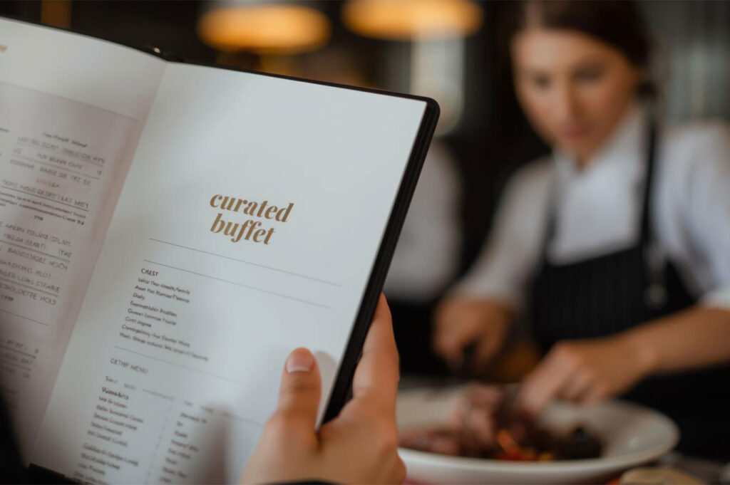 A customer holding an open restaurant menu titled "curated buffet," with a blurred background showing chefs preparing food.