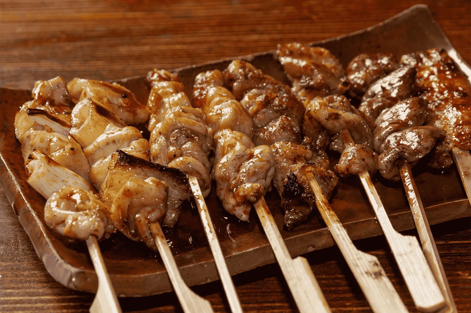 A rectangular ceramic plate filled with assorted Japanese Yakitori, including grilled chicken and leek skewers glazed with savory tare sauce.