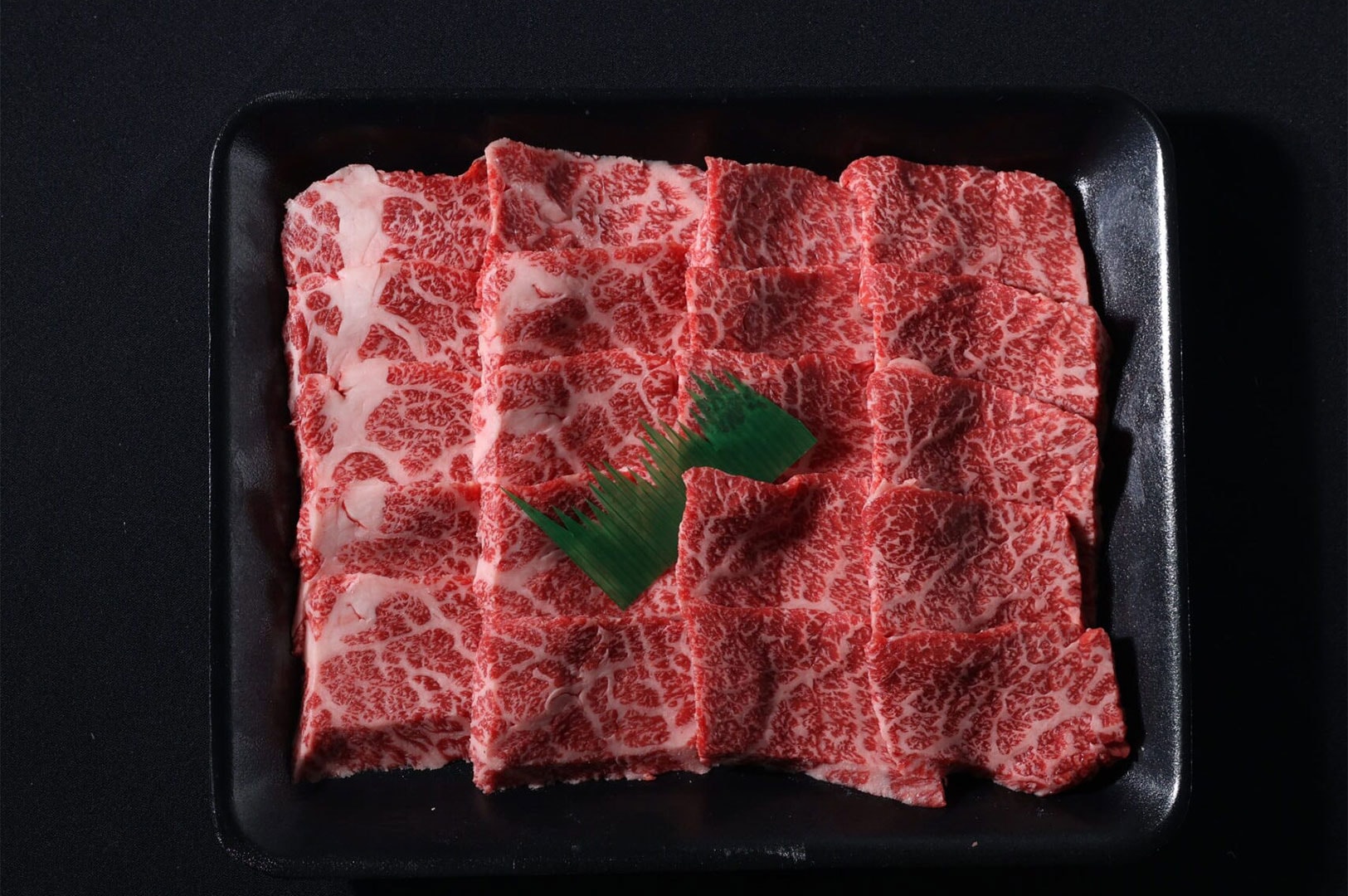 A black serving tray filled with square-cut slices of raw beef featuring intense marbling, prepared for Japanese BBQ grilling.