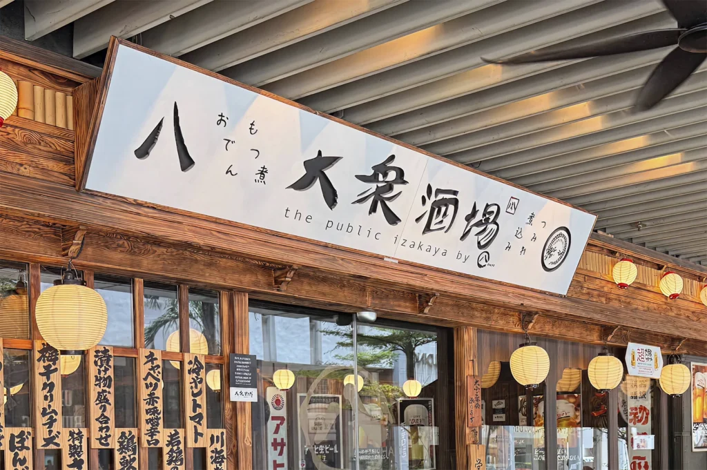 The entrance of "The Public Izakaya" restaurant in Singapore, featuring a prominent sign with Japanese calligraphy, wooden exterior detailing, and traditional paper lanterns hanging along the facade. Various Japanese food items are listed in black script on wooden panels displayed on the glass windows below the sign.
