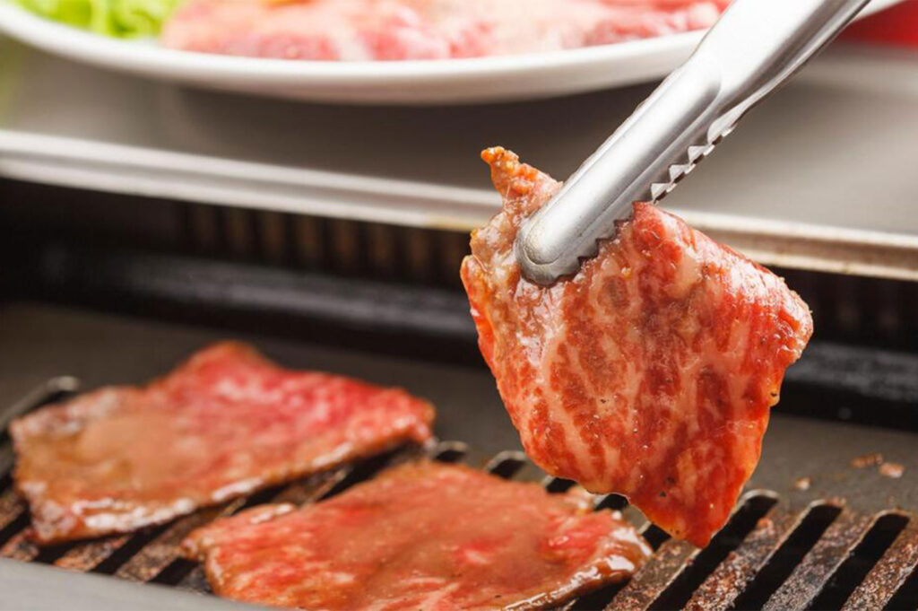 Close-up of metal tongs holding a piece of freshly grilled, juicy Wagyu beef over a hot charcoal grill.