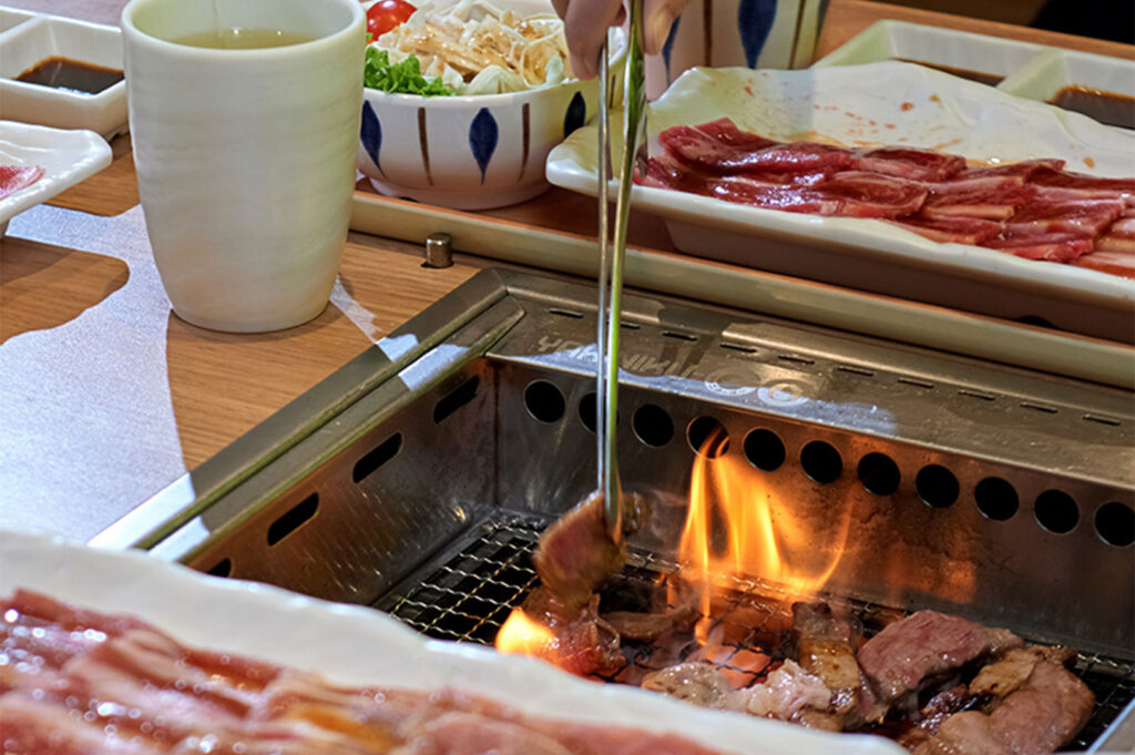 Action shot of tongs placing meat onto a metal Yakiniku grill with flames flaring up, with side dishes and dipping sauces visible on the table.