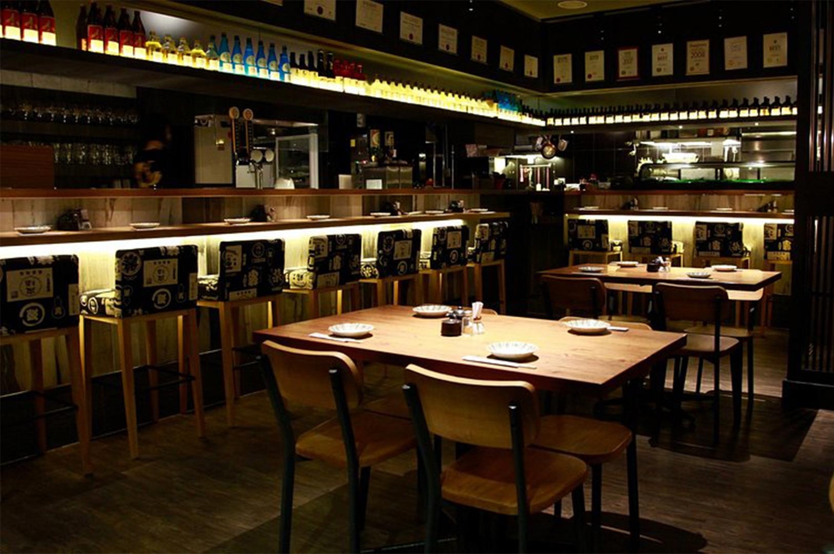 Interior view of a contemporary Japanese restaurant featuring a long wooden bar with stools, ambient under-counter lighting, and neatly arranged dining tables.