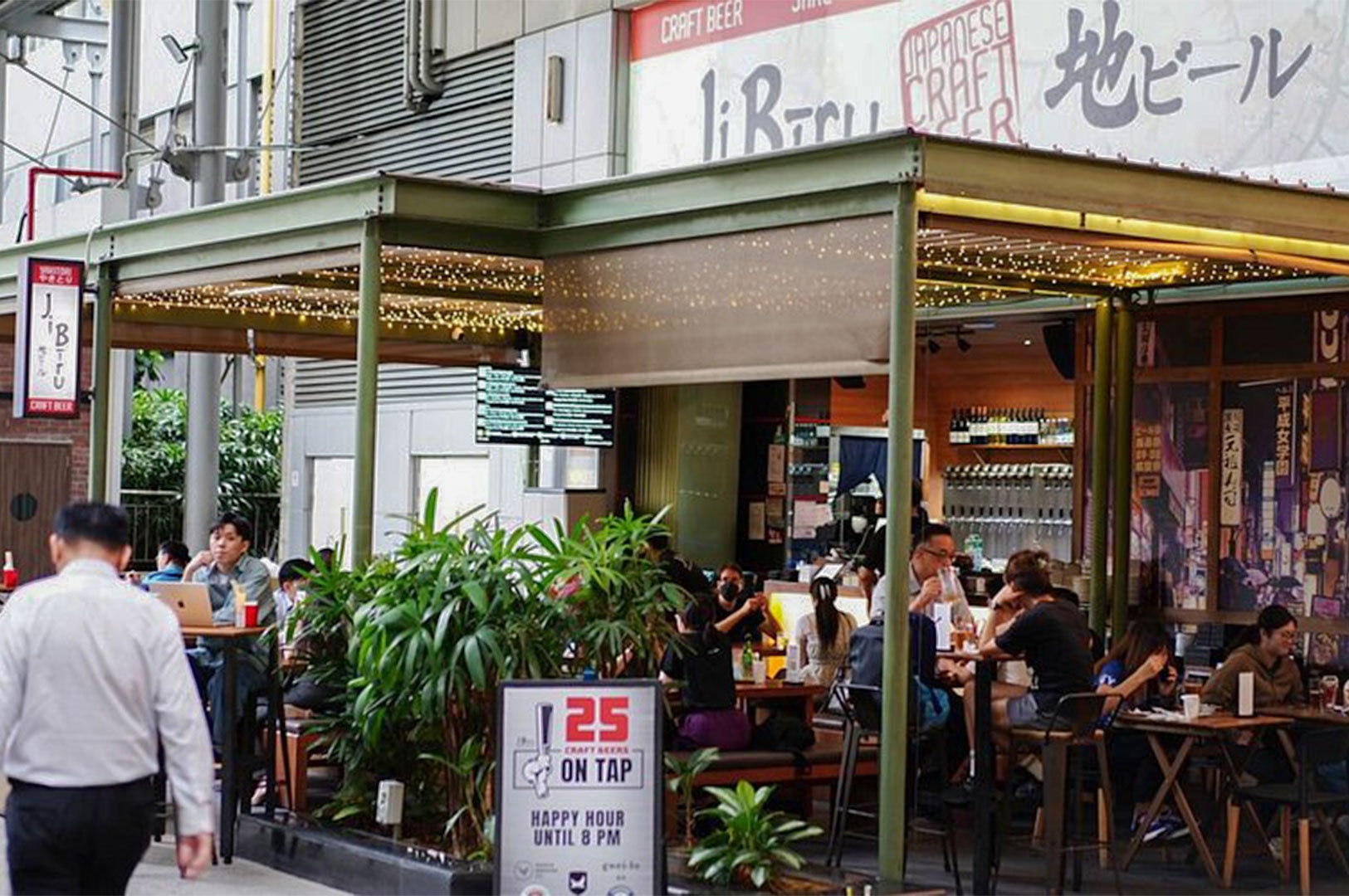 The outdoor alfresco dining area of JiBiru Japanese Craft Beer bar, featuring patrons sitting at wooden tables under string lights in an urban street setting.