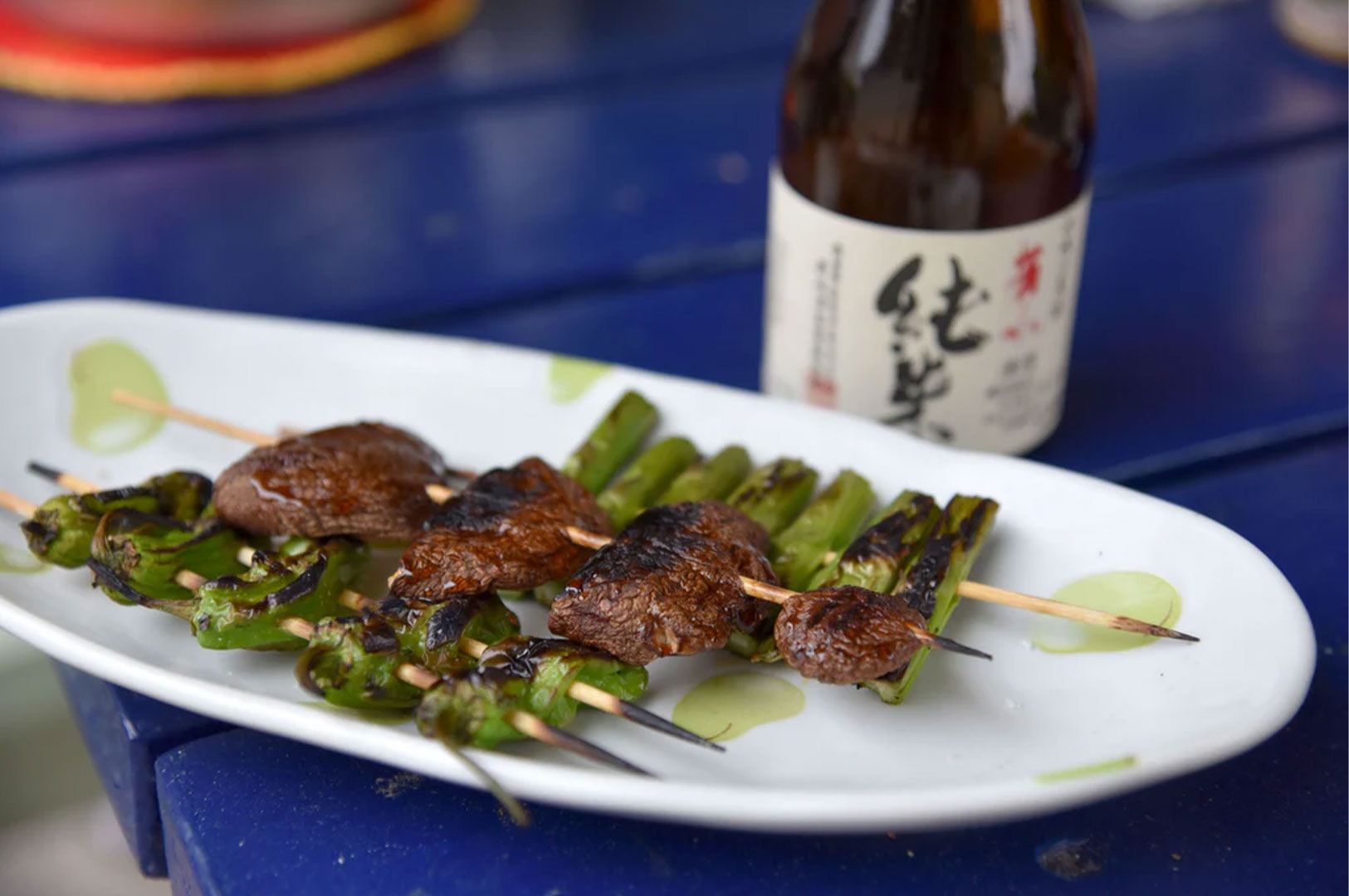 A white plate serving grilled green peppers and meat skewers, accompanied by a traditional bottle of Japanese Sake in the background.