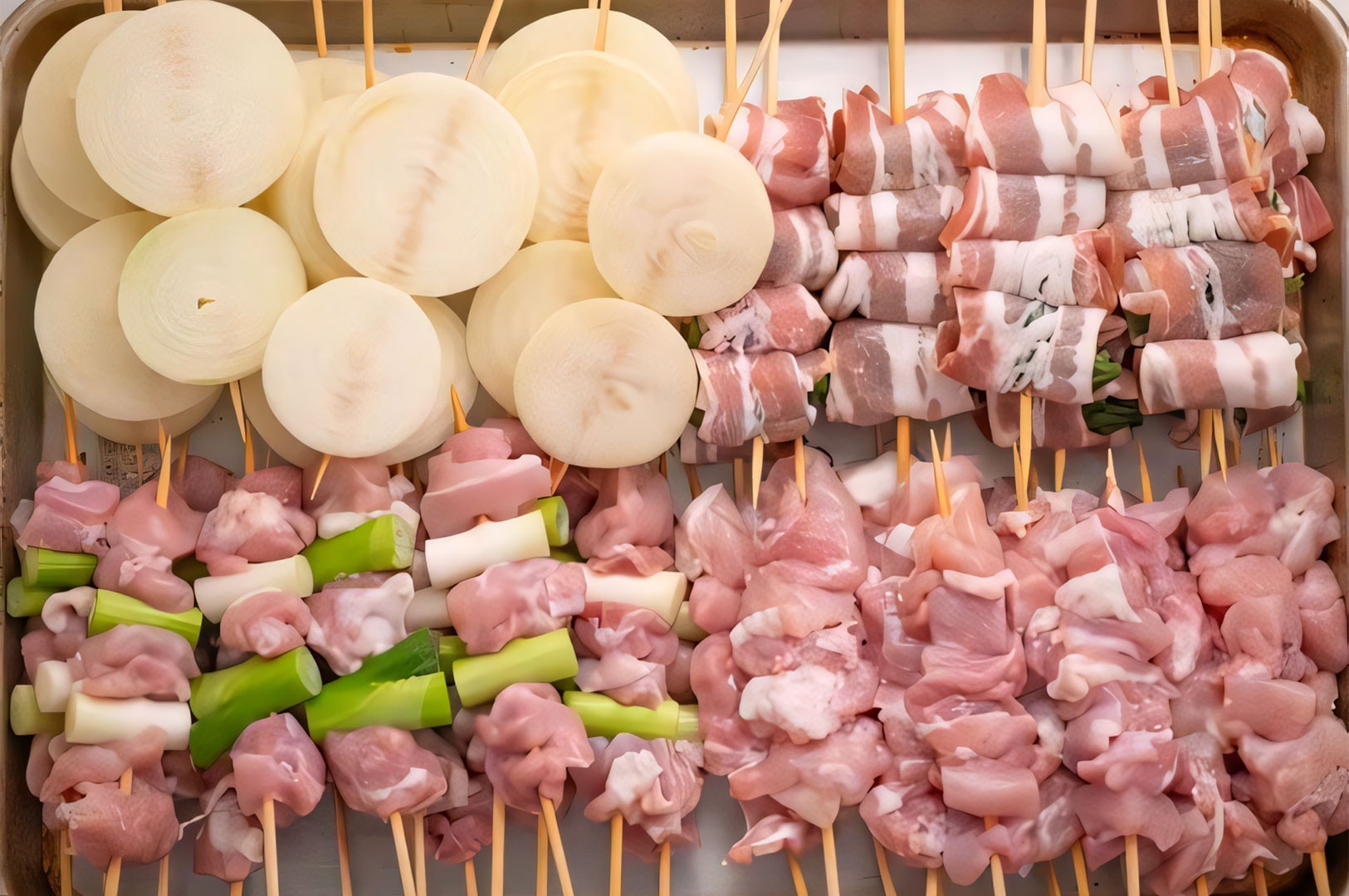A stainless steel tray filled with prepared raw skewers, including sliced onions, bacon-wrapped vegetables, Negima, and chicken meat ready for grilling.