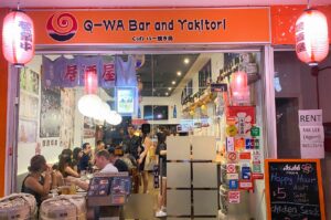 The exterior entrance of Q-WA Bar and Yakitori featuring an orange sign, traditional red paper lanterns, and customers dining inside a cozy Japanese izakaya.