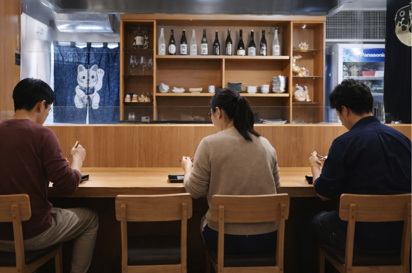 Three people sitting at a minimalist wooden counter in a Japanese restaurant, facing a shelf of sake bottles and a blue "Maneki-neko" (lucky cat) curtain.