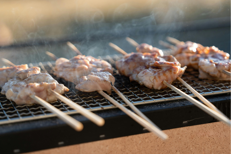 A row of chicken skewers cooking on a wire mesh grill, with wisps of smoke rising against a soft, blurred background.