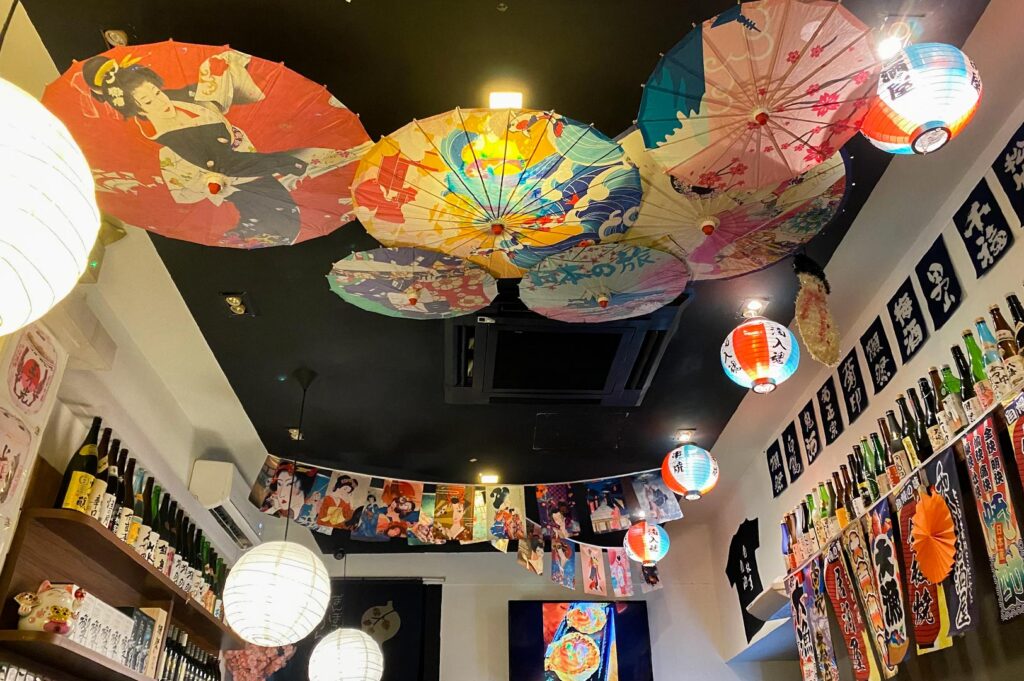 Interior view of Q-WA Bar showing a black ceiling decorated with colorful traditional Japanese wagasa (oil-paper umbrellas), lanterns, and shelves of sake bottles.