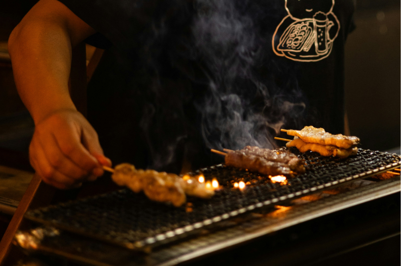 A chef in a black graphic t-shirt grilling various skewers over an open flame, with thick smoke drifting upward.